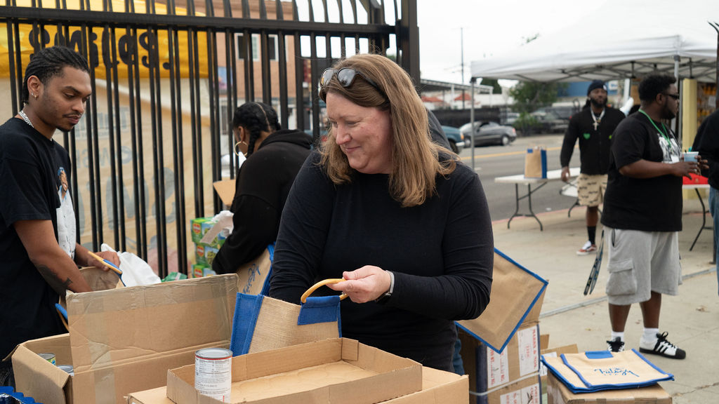 People packing boxes with cans and supplies at an outdoor event near a fence and tent.