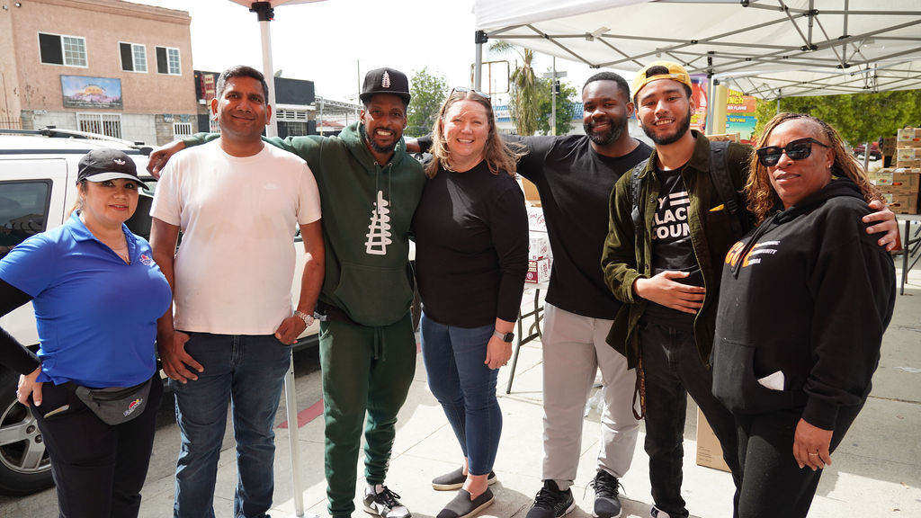 A group of seven people standing together under a tent at an outdoor event, smiling at the camera. There are various buildings and a car in the background.